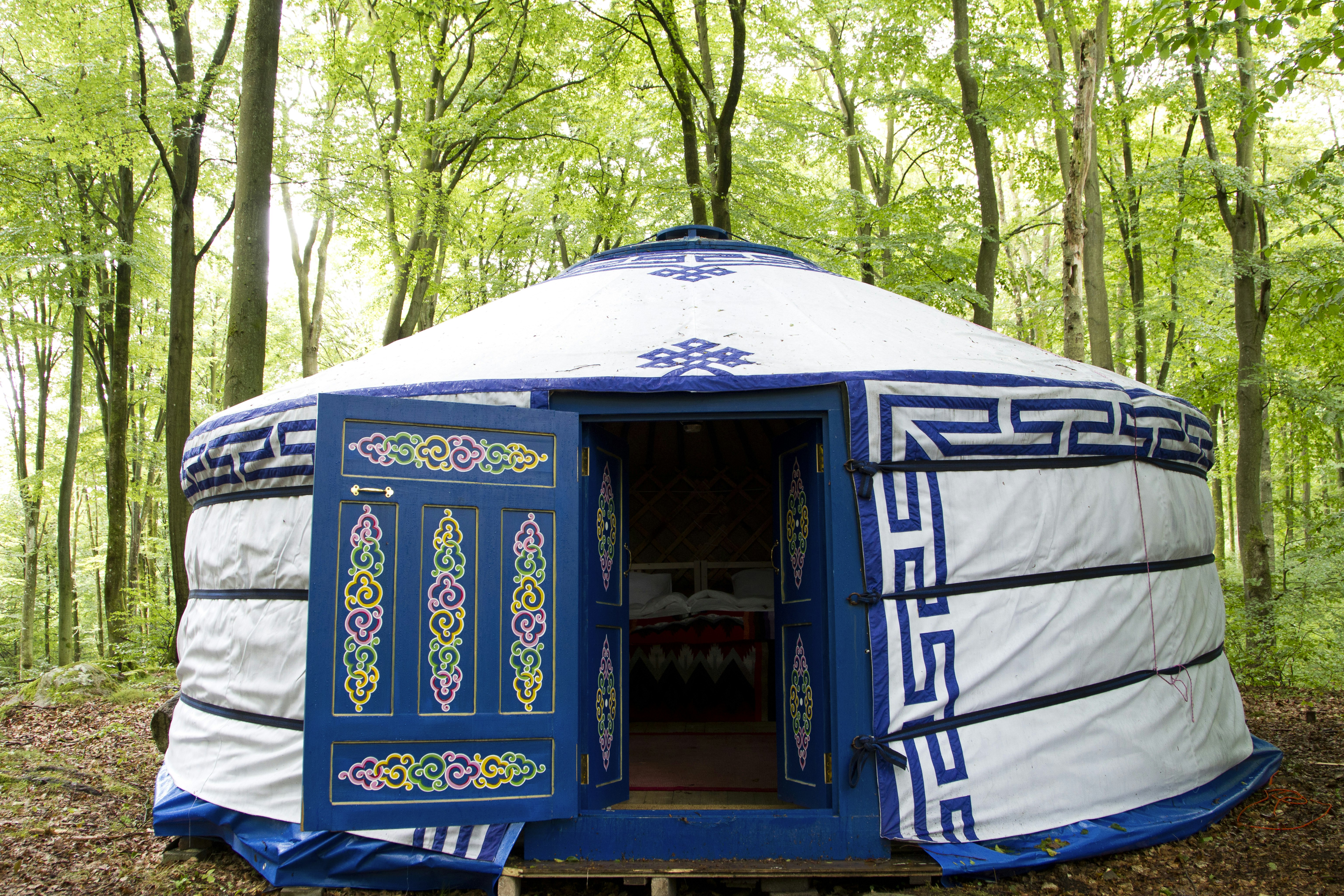 A blue and white wool yurt with geometric blue decorative border around the door and along the top of the yurt, as well as around the peak of the yurt, and a blue door with a pink, yellow, and green intricate knot pattern stands in a bright green forest in Skane, Sweden